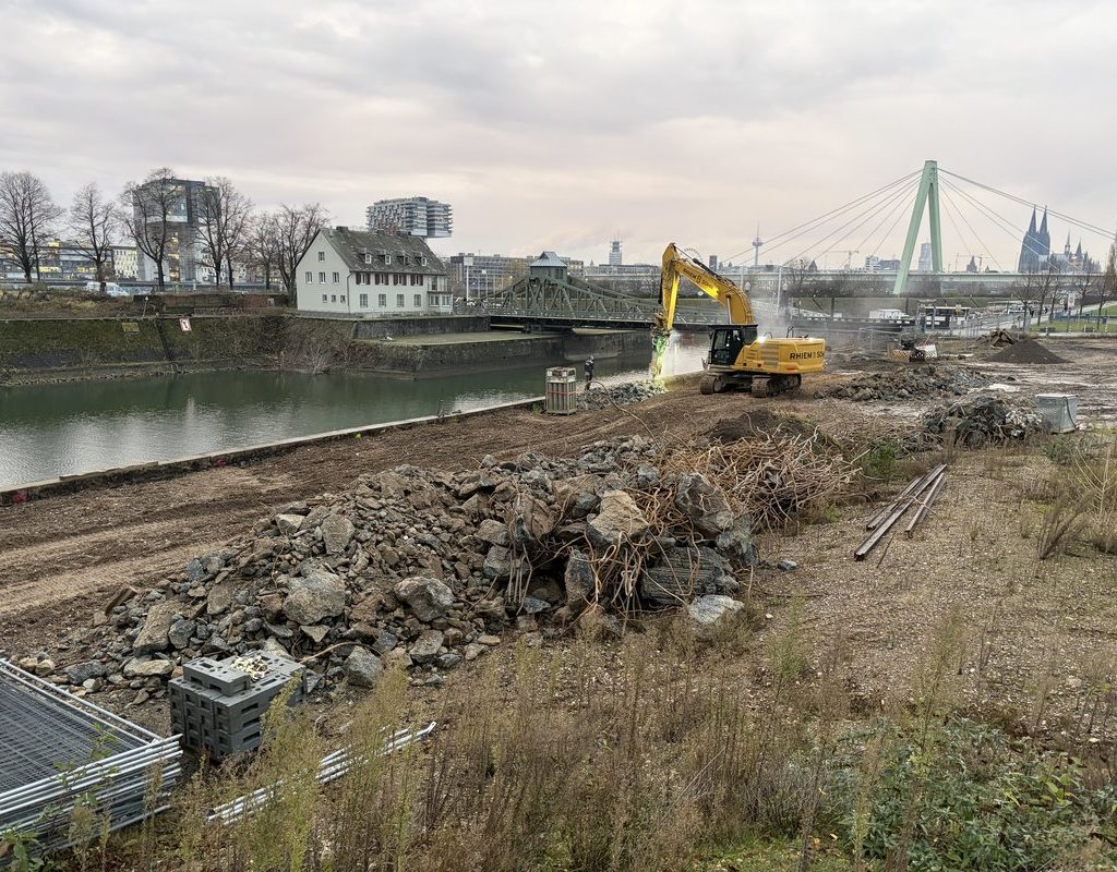 ALT-Text (Kaimauer-Rückbau am Hafenbecken) Abbrucharbeiten an der Kaimauer des Deutzer Hafens in Köln. Ein gelber Kettenbagger bricht die Betonkaimauer am Hafenbecken ab, Staub steigt beim Abbruch auf. Im Vordergrund liegt ein großer Haufen Betonbruchstücke mit freiliegenden Bewehrungsstählen sowie abgelagerte Stahlrohre. Im Hintergrund sind die historische grüne Drehbrücke am Hafeneingang, ein weißes Wohnhaus sowie die Severinsbrücke und der Kölner Dom erkennbar. Links liegt das Hafenbecken, dahinter die Poller Wiese mit kahlen Winterbäumen.