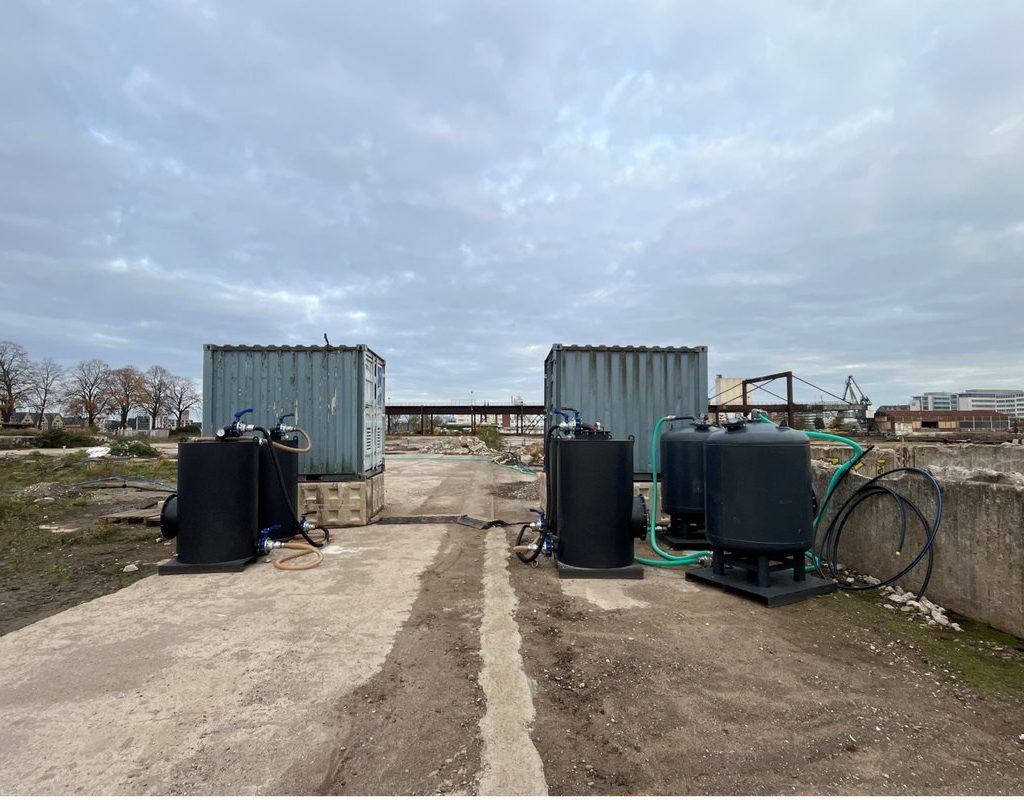 ALT-Text (Air-Sparging-Anlage / Deutzer Hafen) Bodensanierungsanlage auf dem Baugelände des Deutzer Hafens in Köln. Zwei Baucontainer mit je mehreren schwarzen Drucktanks und grünen Schlauchleitungen stehen auf einer Betonplatte — Bestandteile einer Air-Sparging-Anlage zur Grundwassersanierung. Im Hintergrund sind die geräumte Brachfläche, die rostbraune Stahlkonstruktion der alten Verladebrücke sowie der grüne Portalkran am Hafenbecken erkennbar. Bewölkter Winterhimmel über dem Gelände.