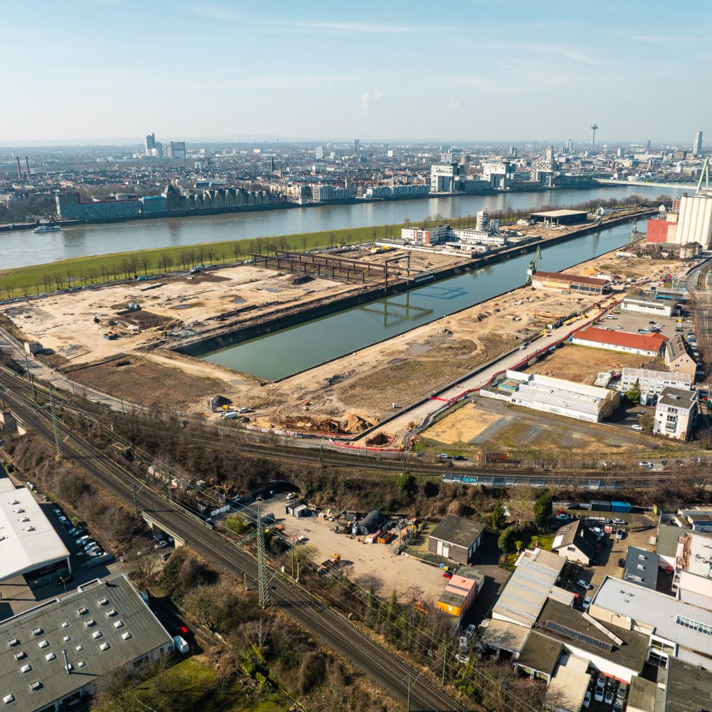 Weiträumige Luftaufnahme des gesamten Deutzer Hafens aus südlicher Perspektive. Das Hafenbecken teilt das Bild. Links das ausgedehnte Baufeld mit dem Portalkran-Kunstwerk. Rechts weitere Bauflächen mit Baucontainern und Kränen. Im Vordergrund Gewerbebebauung, Gleisanlagen und Bahntrassen. Im Hintergrund der Rhein mit Severinsbrücke und der Kölner Skyline mit Dom.